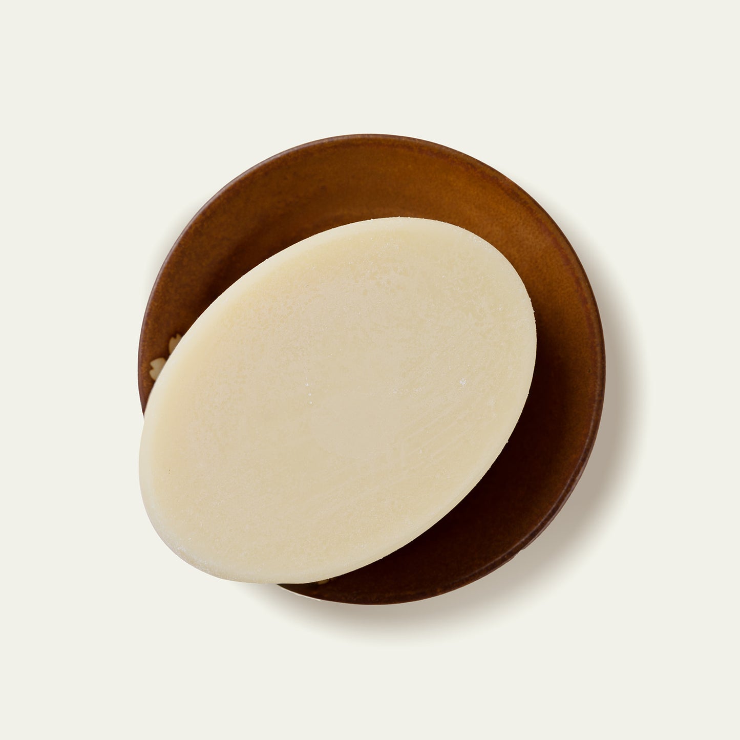 Round M. Carya Body Butter Bar unwrapped on brown dish showing smooth cream surface